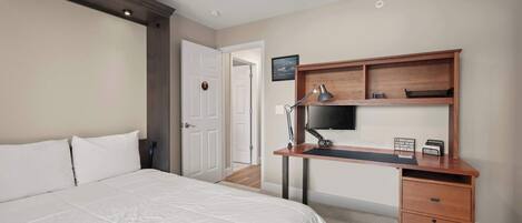 A hotel room with a bed, a desk with a computer monitor, a lamp, and a clock.