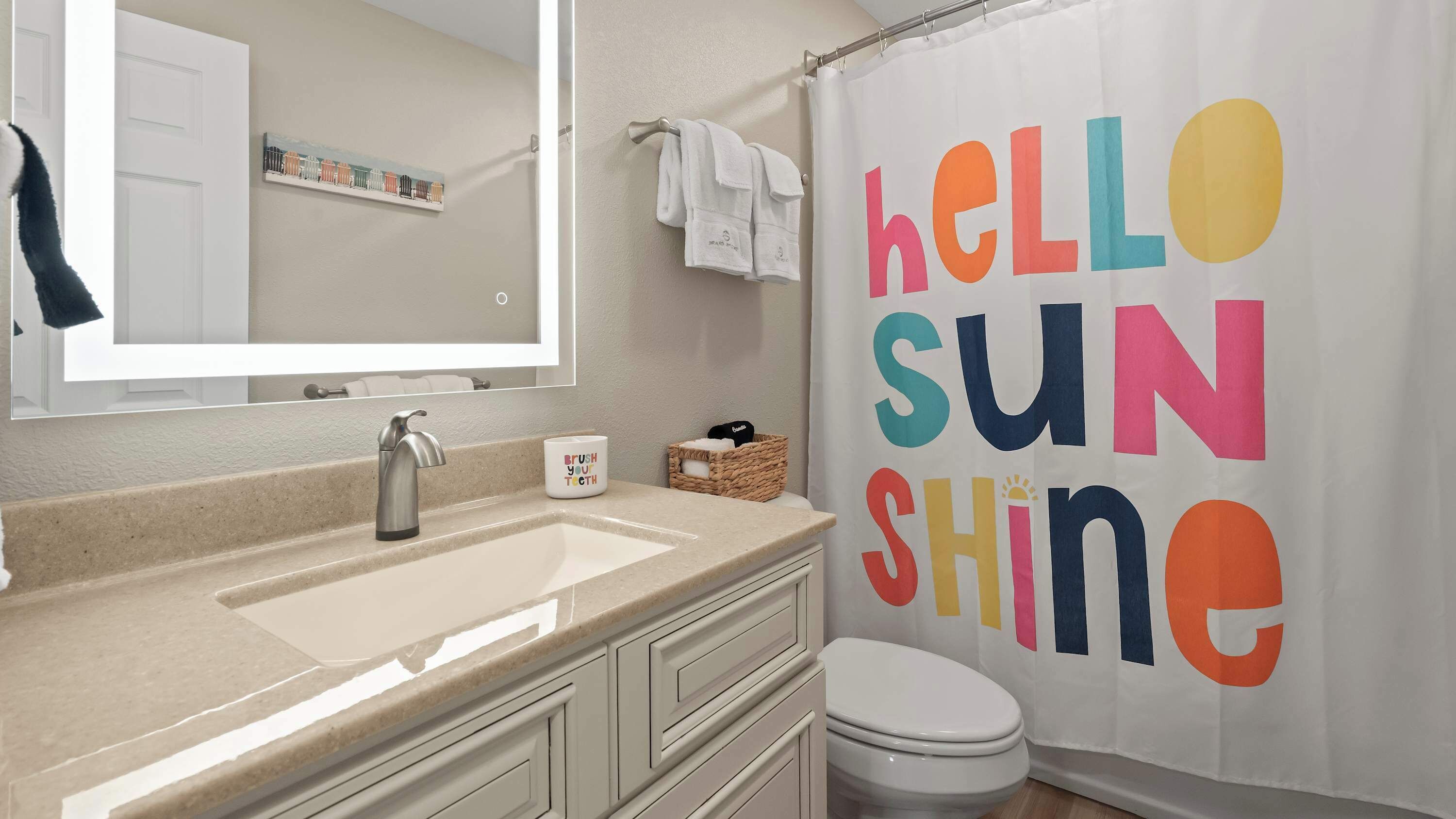 Un baño con un lavamanos color beige, un espejo, un inodoro blanco y una cortina de ducha con la inscripción 'KELLO SUN SHINE'.