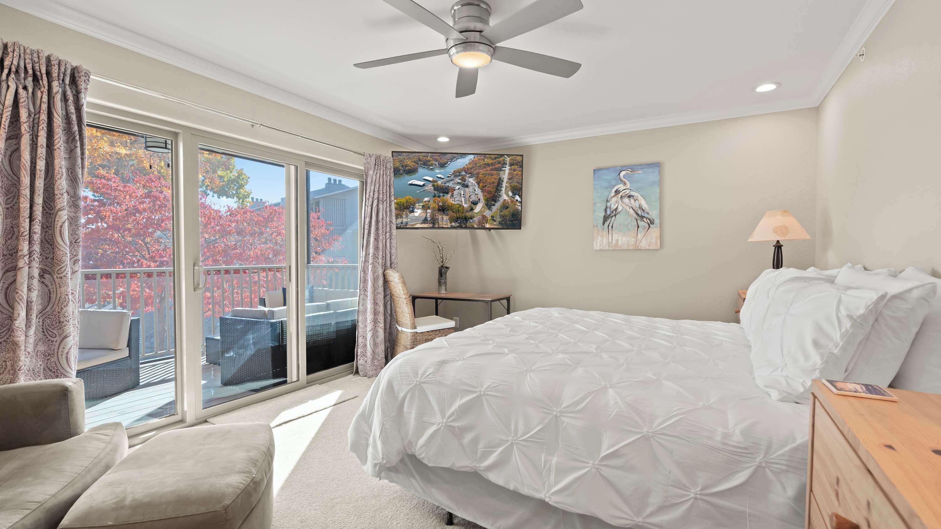 Ein Schlafzimmer mit einem großen Bett, einem Deckenventilator, Blick auf Herbstlaub durch die Glastür und einem Bild an der Wand.
