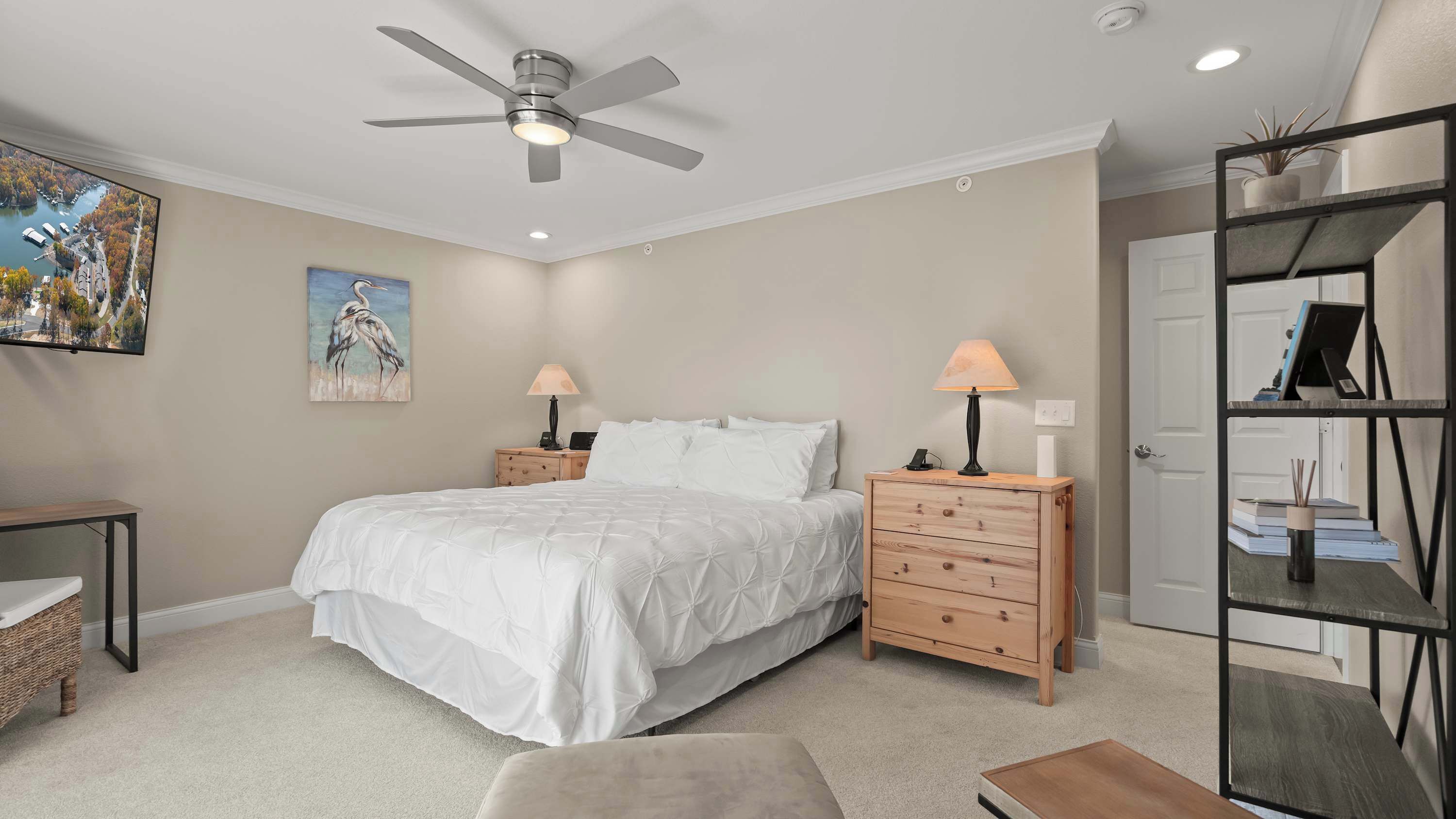 A bedroom with a bed, two bedside tables, a ceiling fan, a painting, a desk, a chair, and a shelving unit.