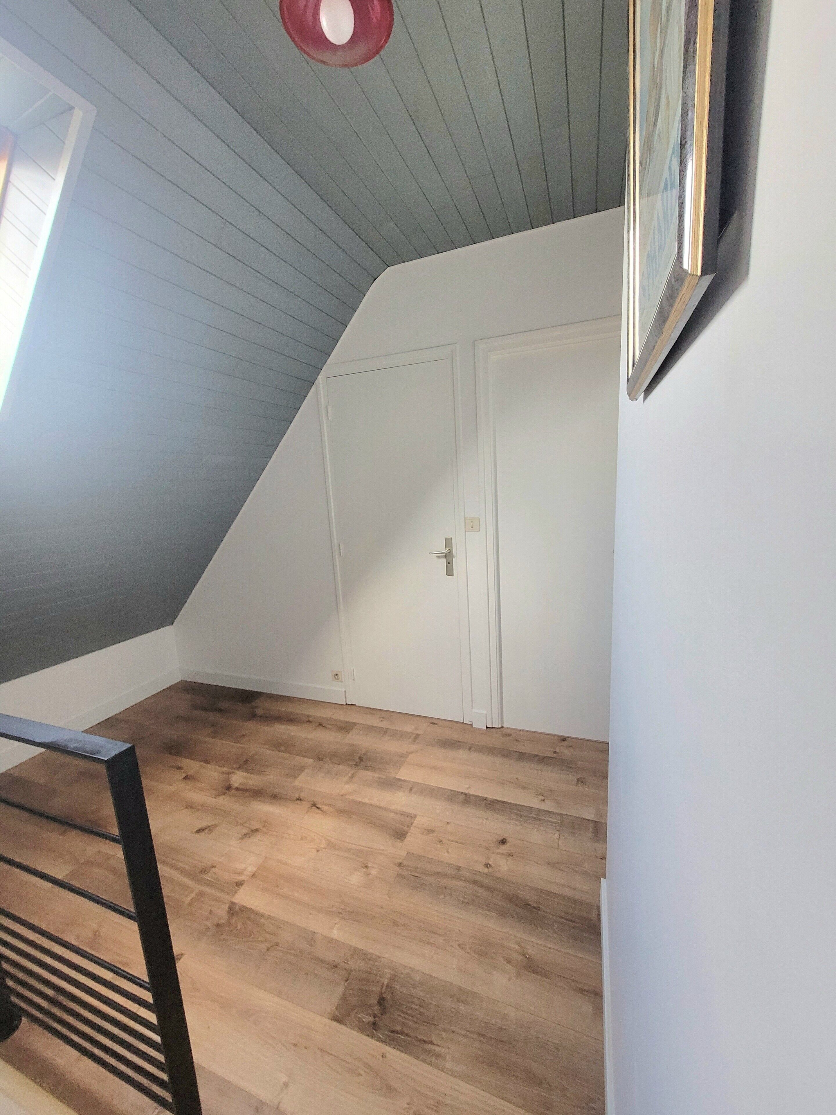 Ein Zimmer mit schrägem Dach, Holzboden, einer weißen Tür und einem gerahmten Bild an der Wand.