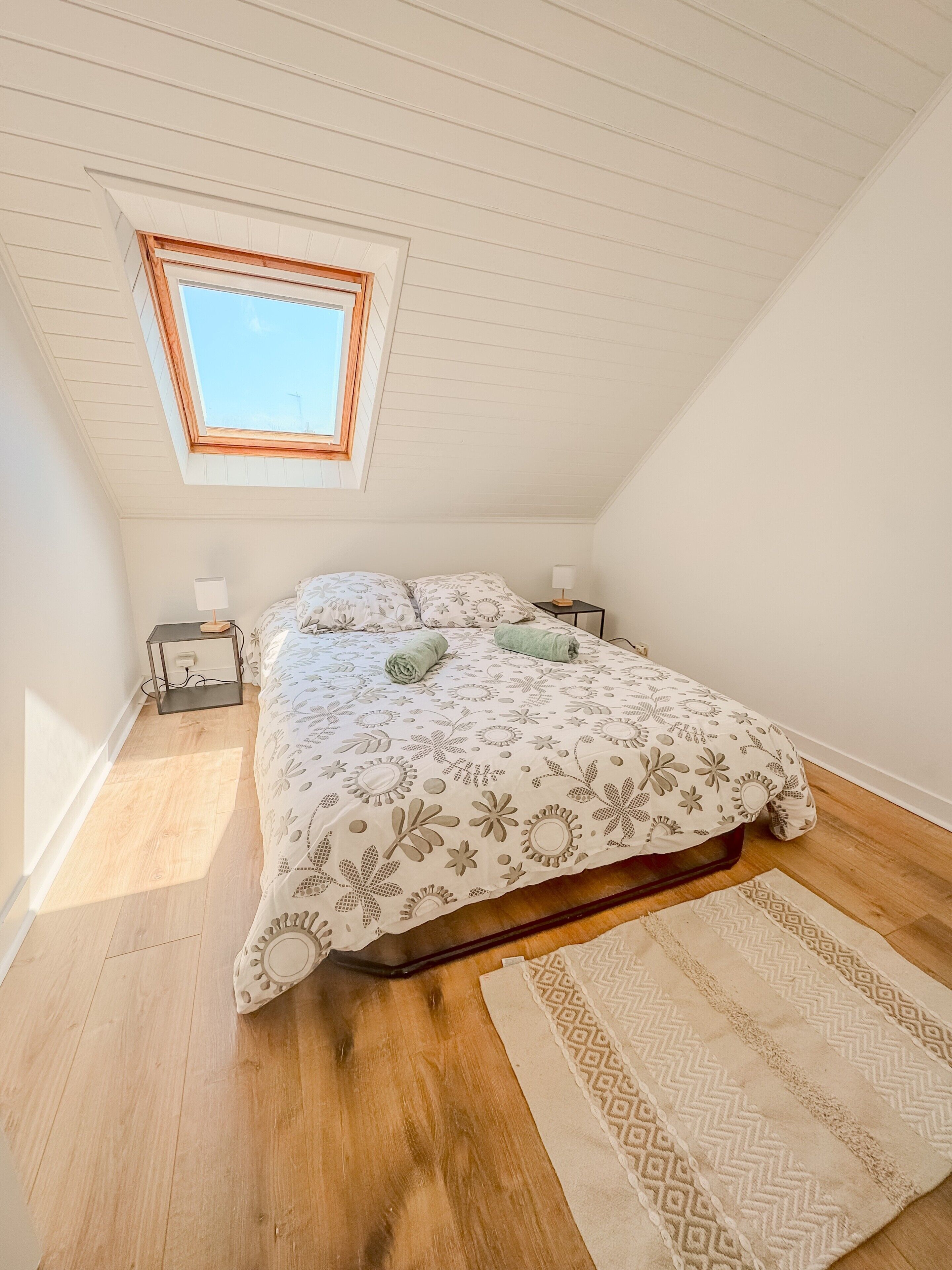 Une chambre avec une lucarne, un lit recouvert d’une literie à motifs, deux tables de chevet avec des lampes et un tapis sur le sol.