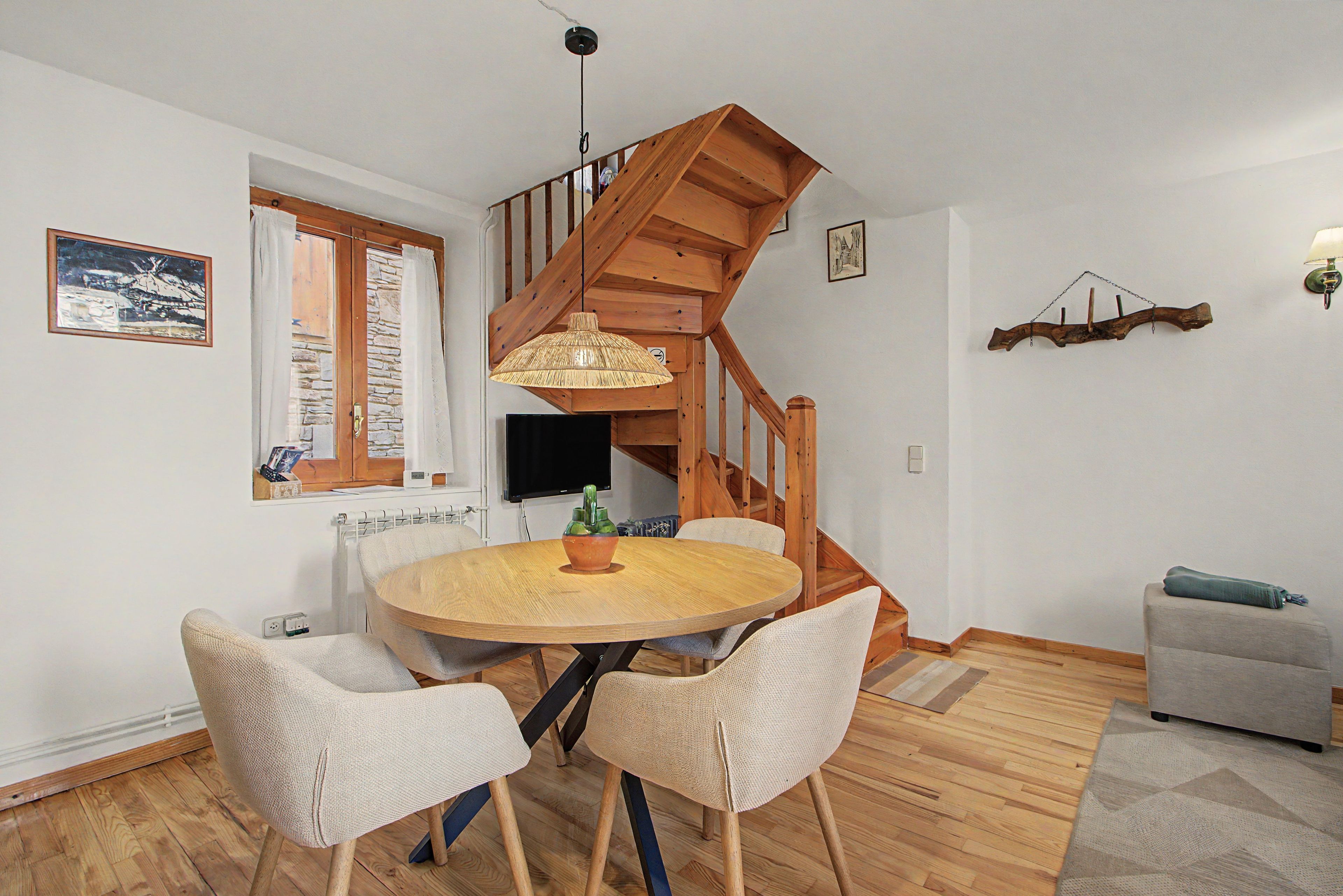 Een eethoek met een ronde houten tafel, twee stoelen, een hanglamp, een trap en een televisie.