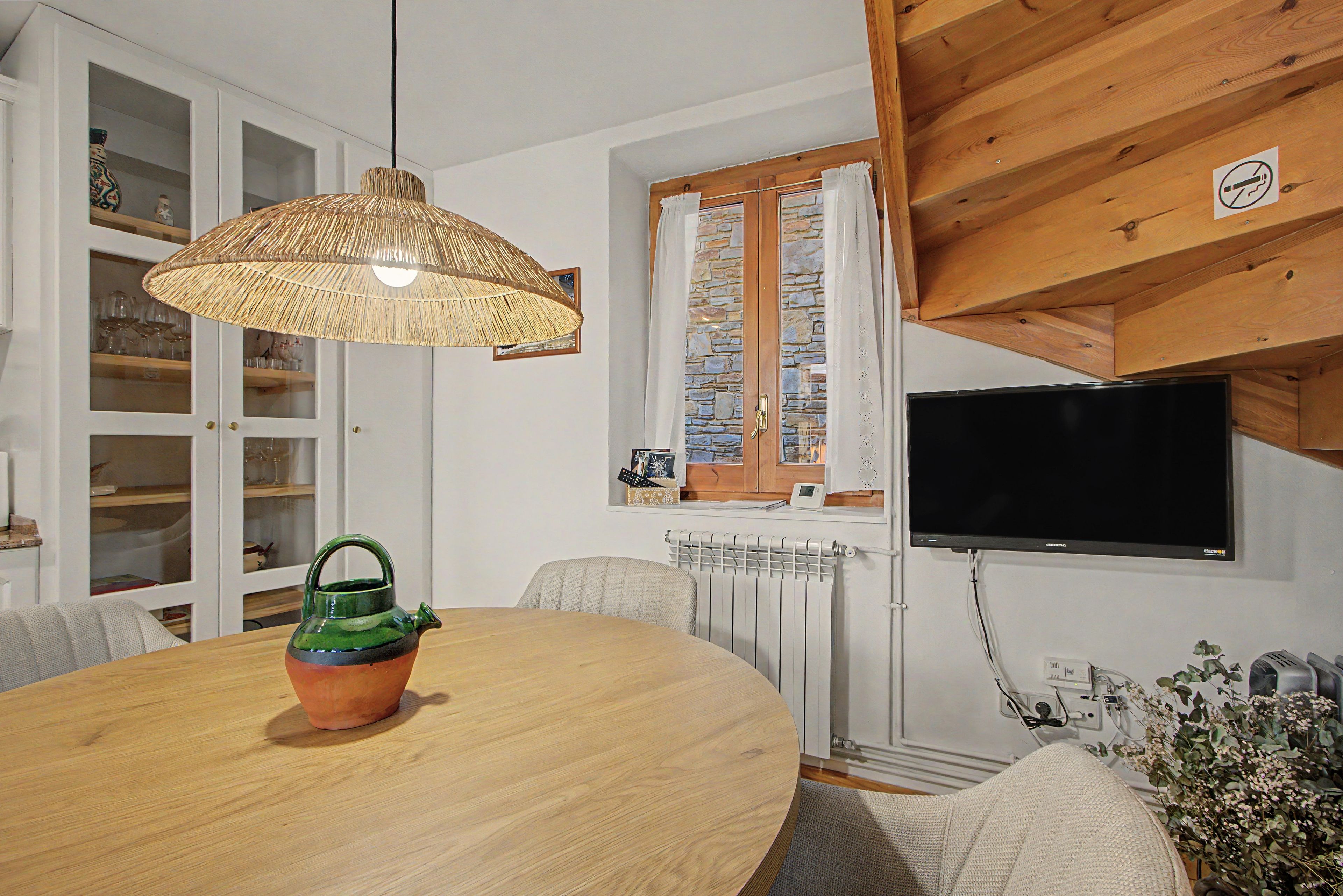 Een eetgedeelte met een ronde tafel, een rieten lamp, een televisie en een raam met gordijnen.