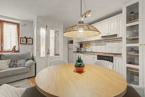 Spiseplads med et rundt træbord, en vævet pendel og et køkken i baggrunden.