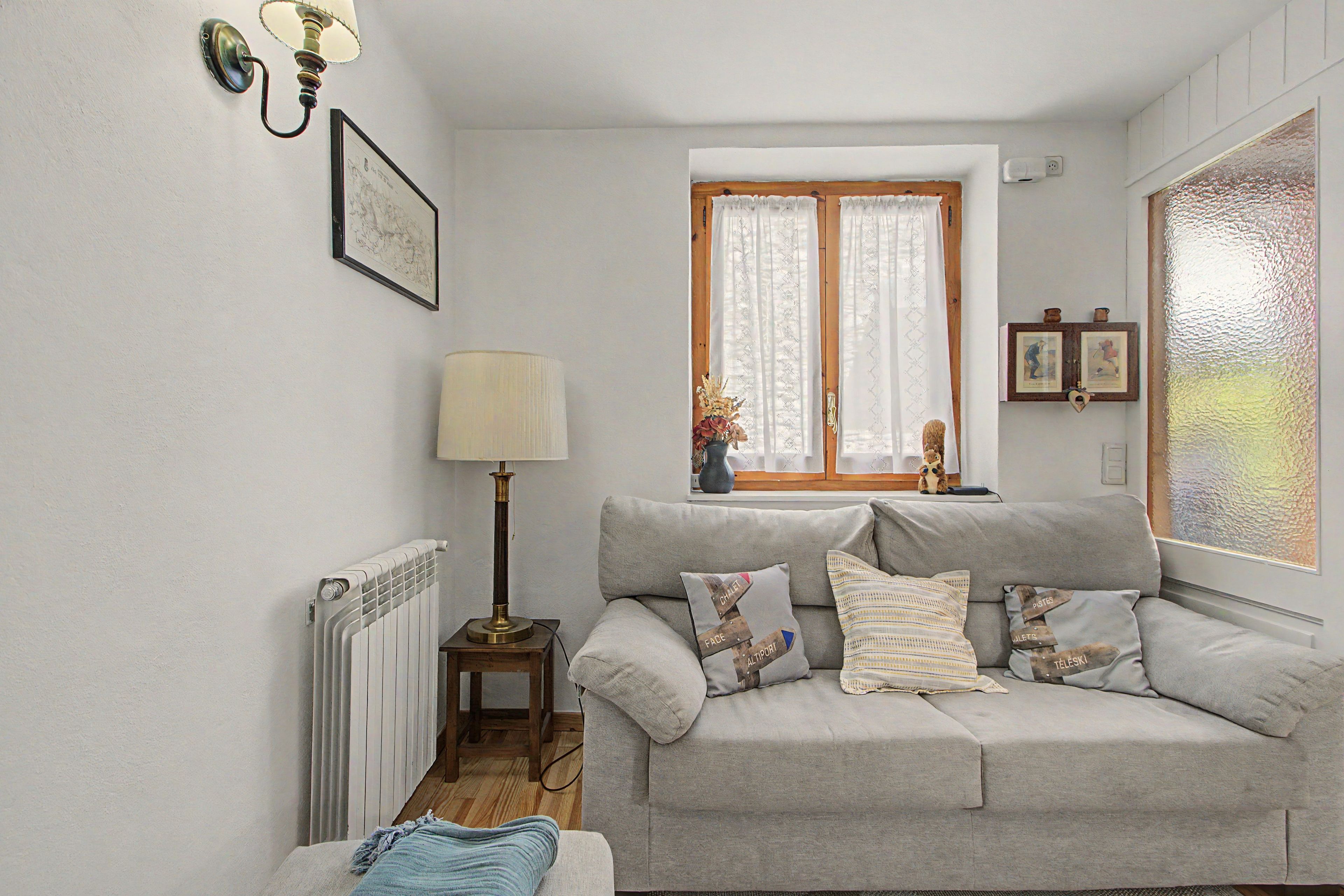 Een knus woongedeelte met een grijze bank, een bijzettafeltje met een lamp, een radiator en een raam met gordijnen.