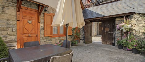 Un bâtiment en pierre avec une porte et des volets en bois, une table avec des chaises et des plantes en pot.