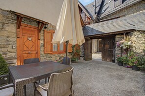 A stone building with a wooden door and window shutters, a table with chairs, and potted plants.