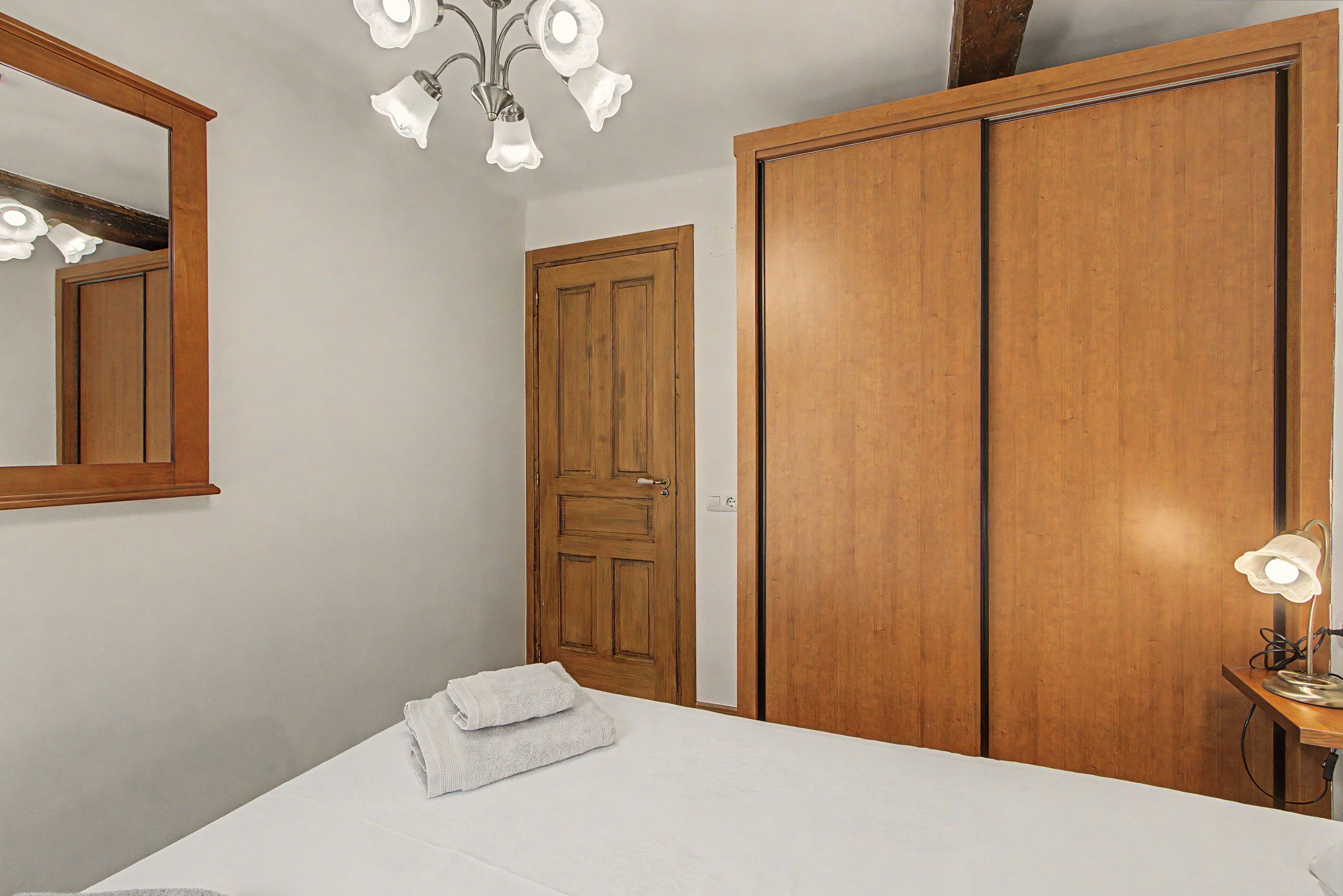 Een slaapkamer met een houten garderobe, een bed met een witte spreideken, een badkamer met een houten deur en een spiegel aan de muur.