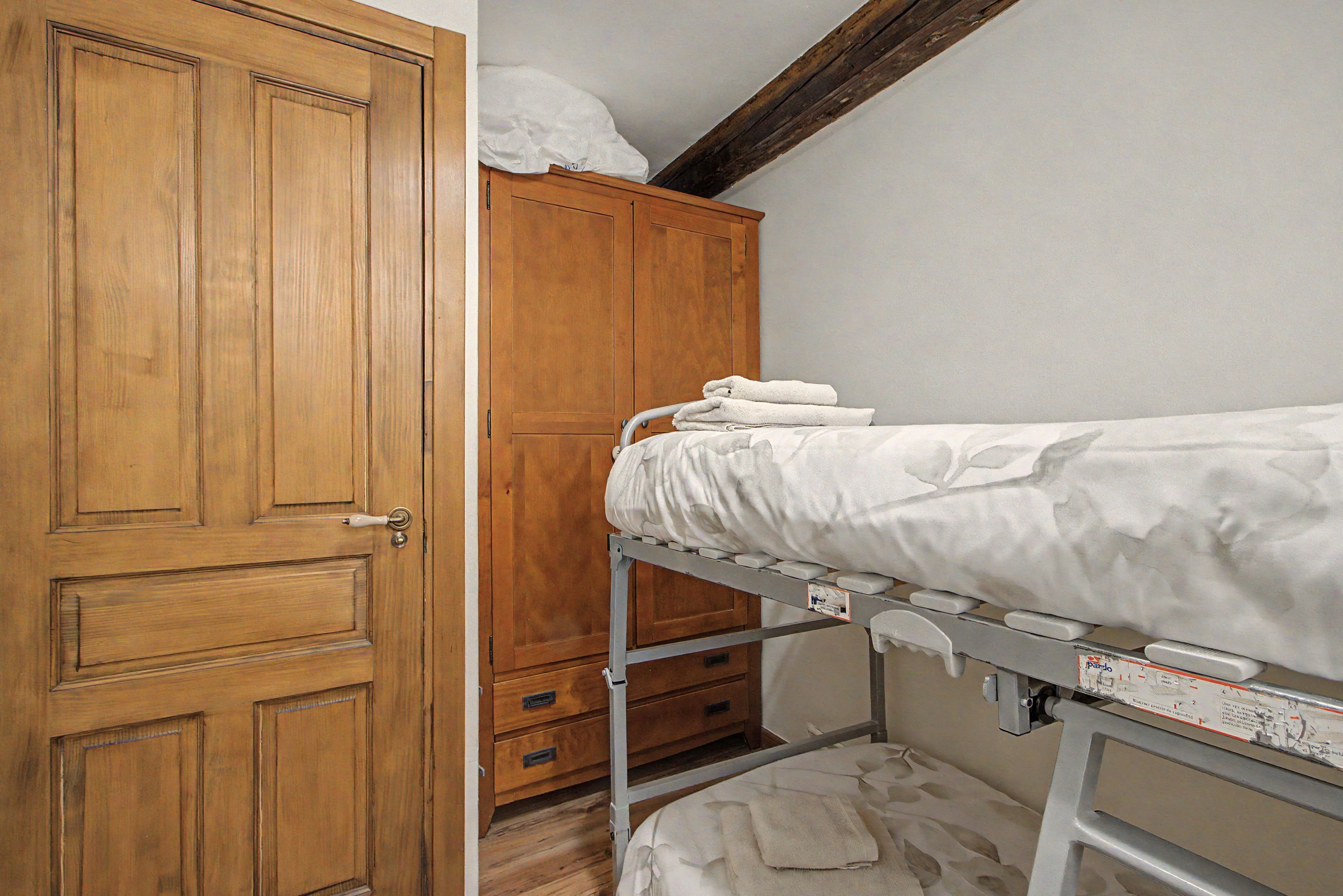 A room with a wooden wardrobe, a metal bunk bed with a mattress, and a small wooden cabinet.