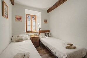 Une petite chambre à coucher accueillante, avec deux lits simples, une table de chevet en bois avec une lampe, une fenêtre donnant sur un beau paysage et des œuvres d’art accrochées au mur.