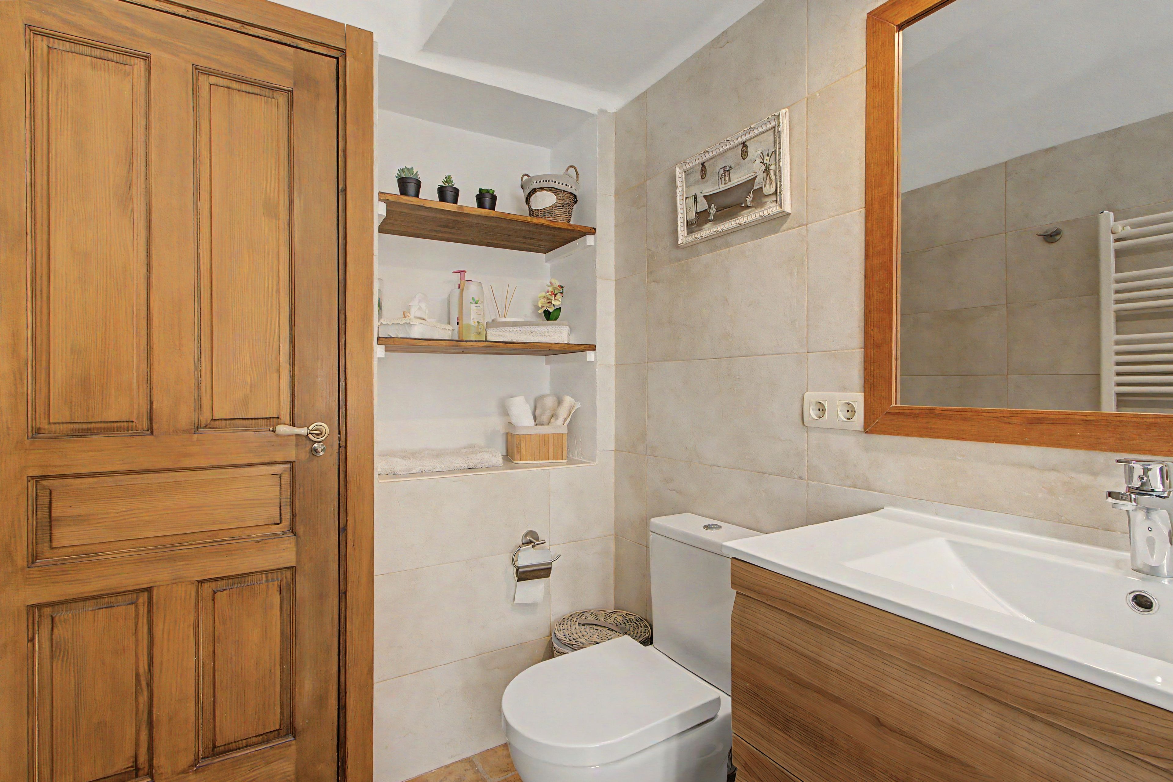 Un baño con puerta de madera, inodoro blanco, lavamanos con espejo y repisas con artículos de aseo.