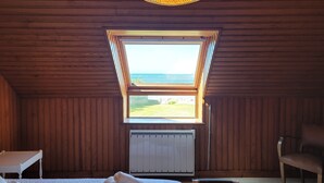 Ein Zimmer mit Holzwänden, einer schrägen Decke, einem großen Fenster mit Meerblick, einem Bett mit zusammengelegtem Handtuch, einem Stuhl und einem Tisch.