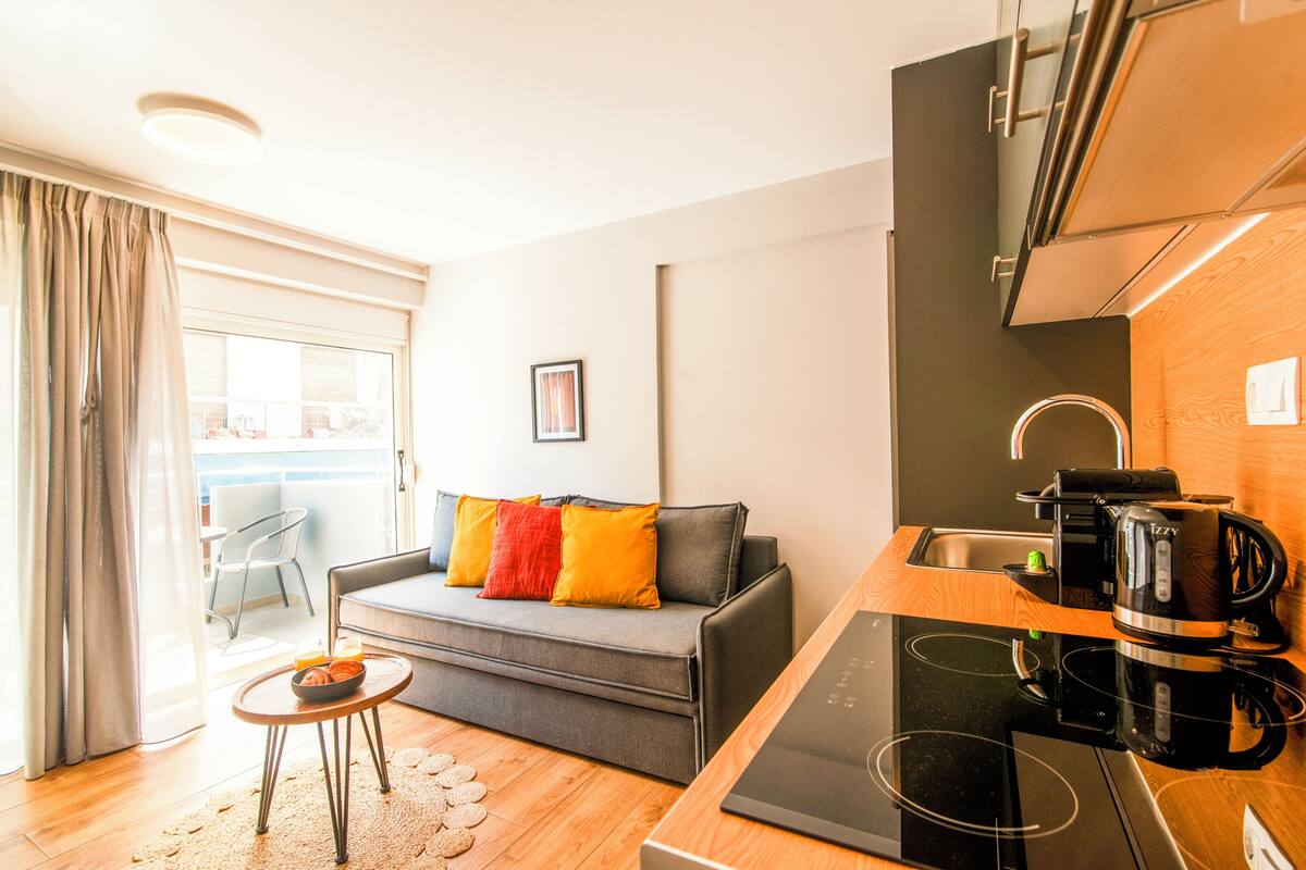 Μια σύγχρονη μικρή κουζίνα με μια μαύρη κουζίνα, μια καφετιέρα και έναν νεροχύτη. Υπάρχει ένας γκρι καναπές με πολύχρωμα μαξιλάρια και ένα μικρό στρογγυλό τραπέζι με ένα μπολ με φρούτα. Στο βάθος διακρίνεται μια καρέκλα και στα αριστερά υπάρχει ένα παράθυρο με κουρτίνες.