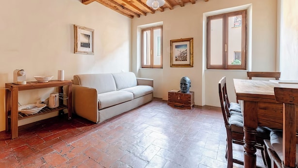 A cozy living room with a sofa, wooden table, and chairs. There are two windows and a ceiling with exposed beams. - Apartment in the Historic Center of Pietrasanta (Pietrasanta)