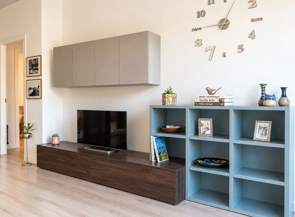 A modern living room with a flat-screen TV, a wooden entertainment unit, a light blue shelving unit with decorative items, and a wall clock.