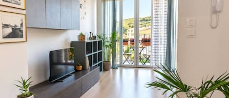 A modern living room with a flat-screen TV, dark wood furniture, a potted plant, and a view of a balcony with outdoor furniture.