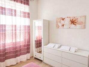 Ein Schlafzimmer mit Bett, Kleiderschrank, Spiegel, einem Fenster mit Vorhängen und einem Wandbild.