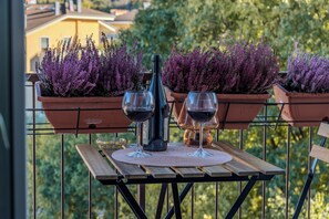Ein Holztisch mit zwei Weingläsern und einer Flasche auf einem Balkon mit violetten Topfpflanzen.