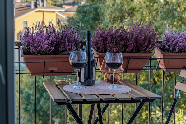 Uma mesa de madeira com duas taças de vinho e uma garrafa numa varanda com plantas roxas em vasos.