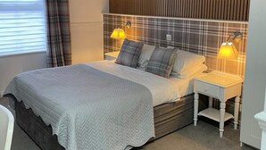 A bedroom with a bed, two bedside lamps, a nightstand, a window with curtains, and a wooden panel wall. - Hunters Meet Ltd  (Hatfield Heath)
