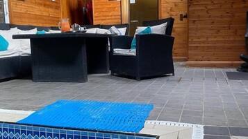A wooden cabin with a pool area, furnished with black wicker sofas and a blue mat.
