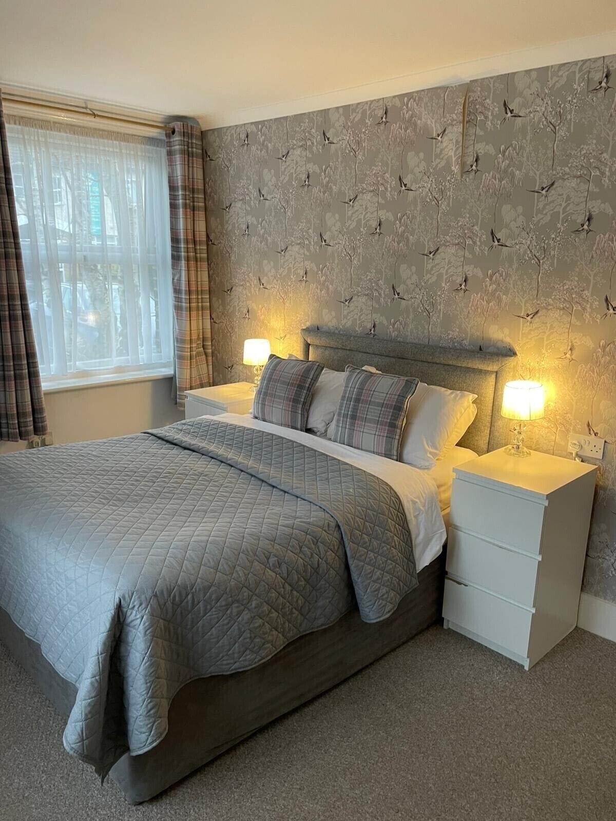 Un dormitorio con una cama grande, dos mesitas de noche con lámparas, una ventana con cortinas y una pared decorada con un estampado de pájaros.