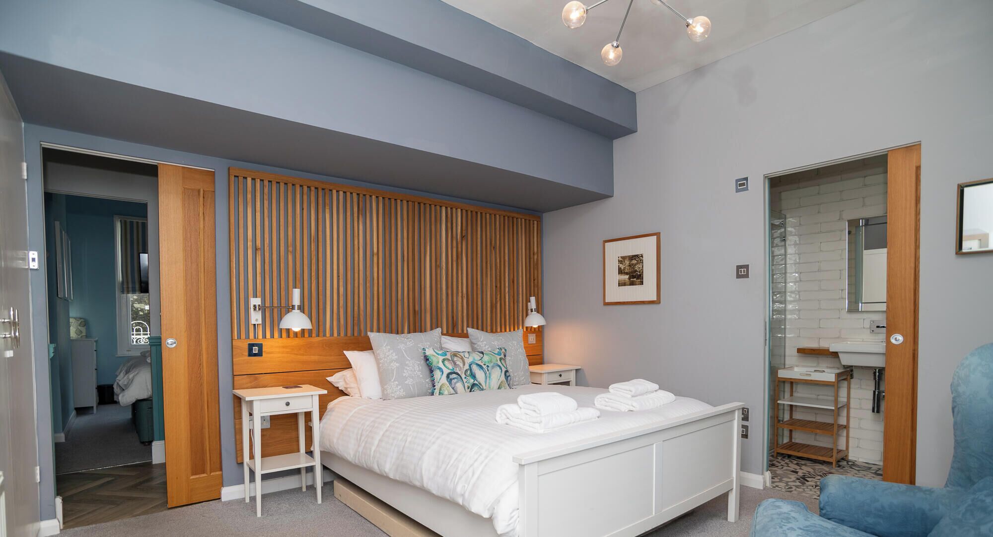 Un dormitorio con cama, mesitas de noche, cabecera de madera, un cuadro enmarco y un baño con lavamanos y espejo.