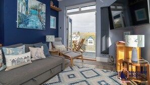 Una sala acogedora con un sofá gris, una mesa de centro de madera y un tapete azul estampado. Un cuadro grande decora la pared, y una ventana ofrece vistas al exterior.