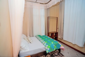 Un dormitorio con cama, mesita de noche, espejo y ventana con cortinas traslúcidas.