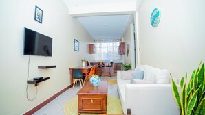 A modern living room with a flat-screen TV, a dining area with a table and chairs, and a kitchen in the background. - Volunteer Hostel (Moshi)