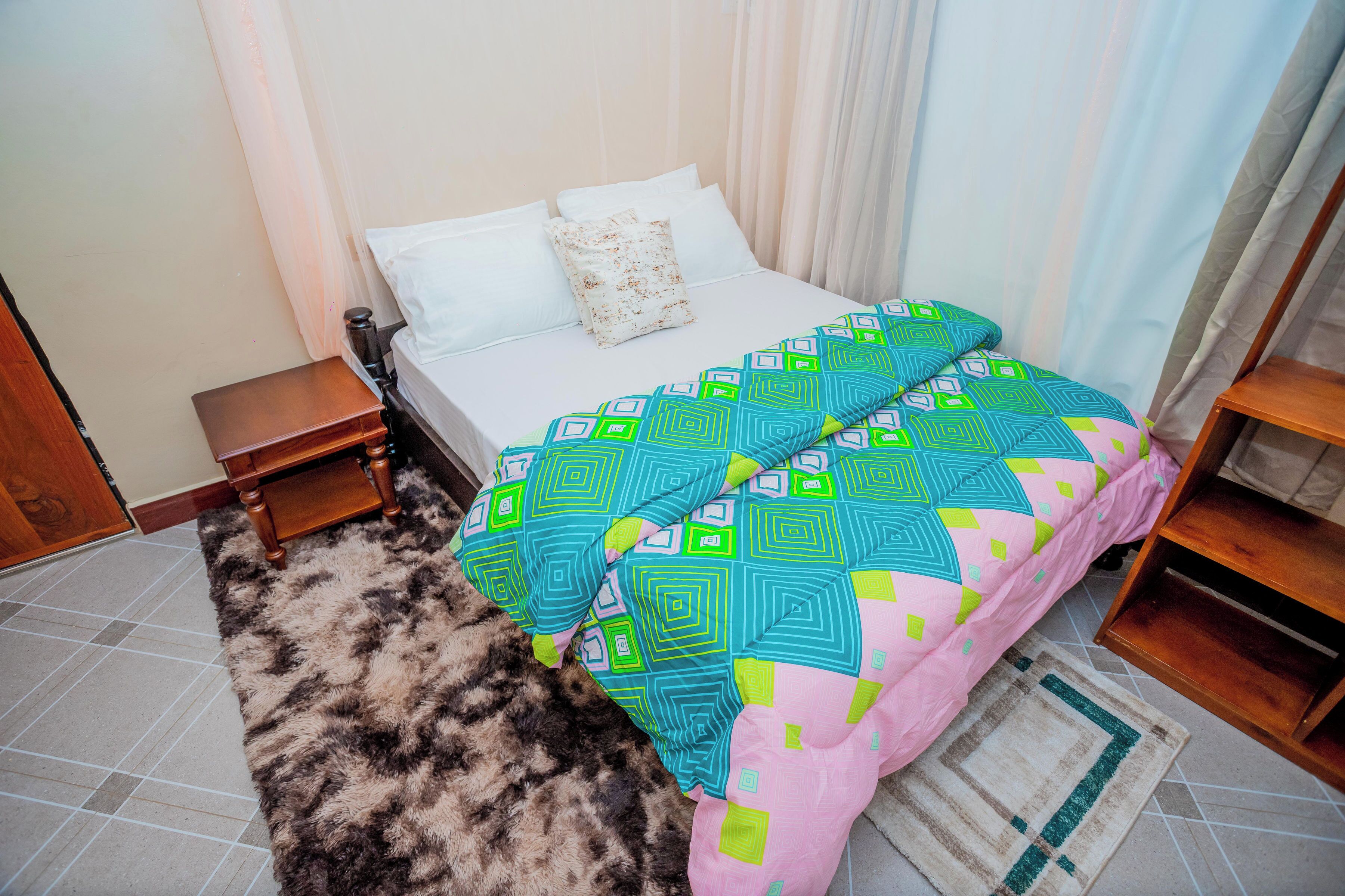 A bedroom with a bed, a nightstand, a wooden chair, a patterned rug, and a window with curtains.