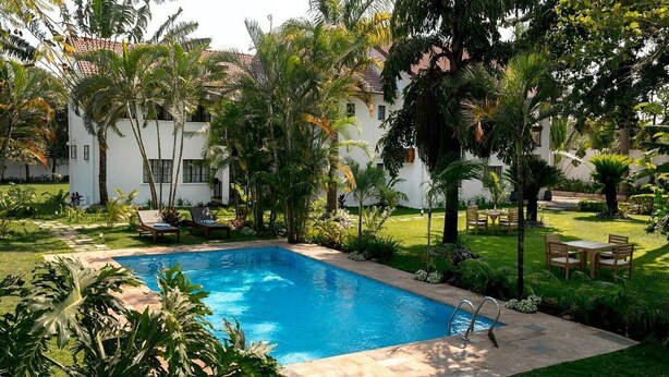 A swimming pool surrounded by lush greenery and palm trees, with a patio area and outdoor furniture. - Volunteer Hostel (Moshi)