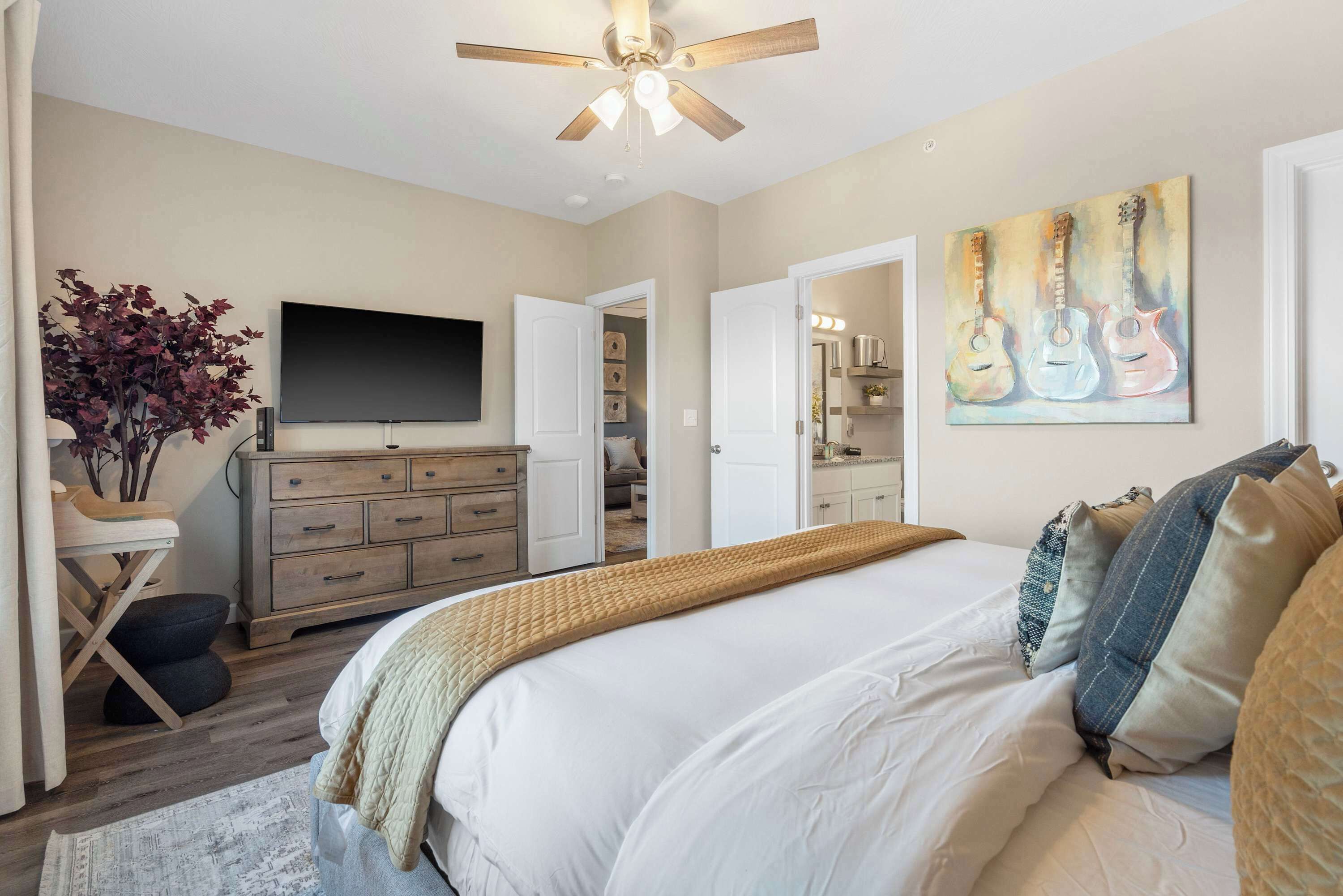 A bedroom with a bed, a ceiling fan, a TV, a dresser, a plant, and a painting of guitars.