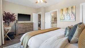 A bedroom with a bed, a ceiling fan, a TV, a dresser, a plant, and a painting of guitars. - Crossroads Vistas - 3bdr Condo (Branson)