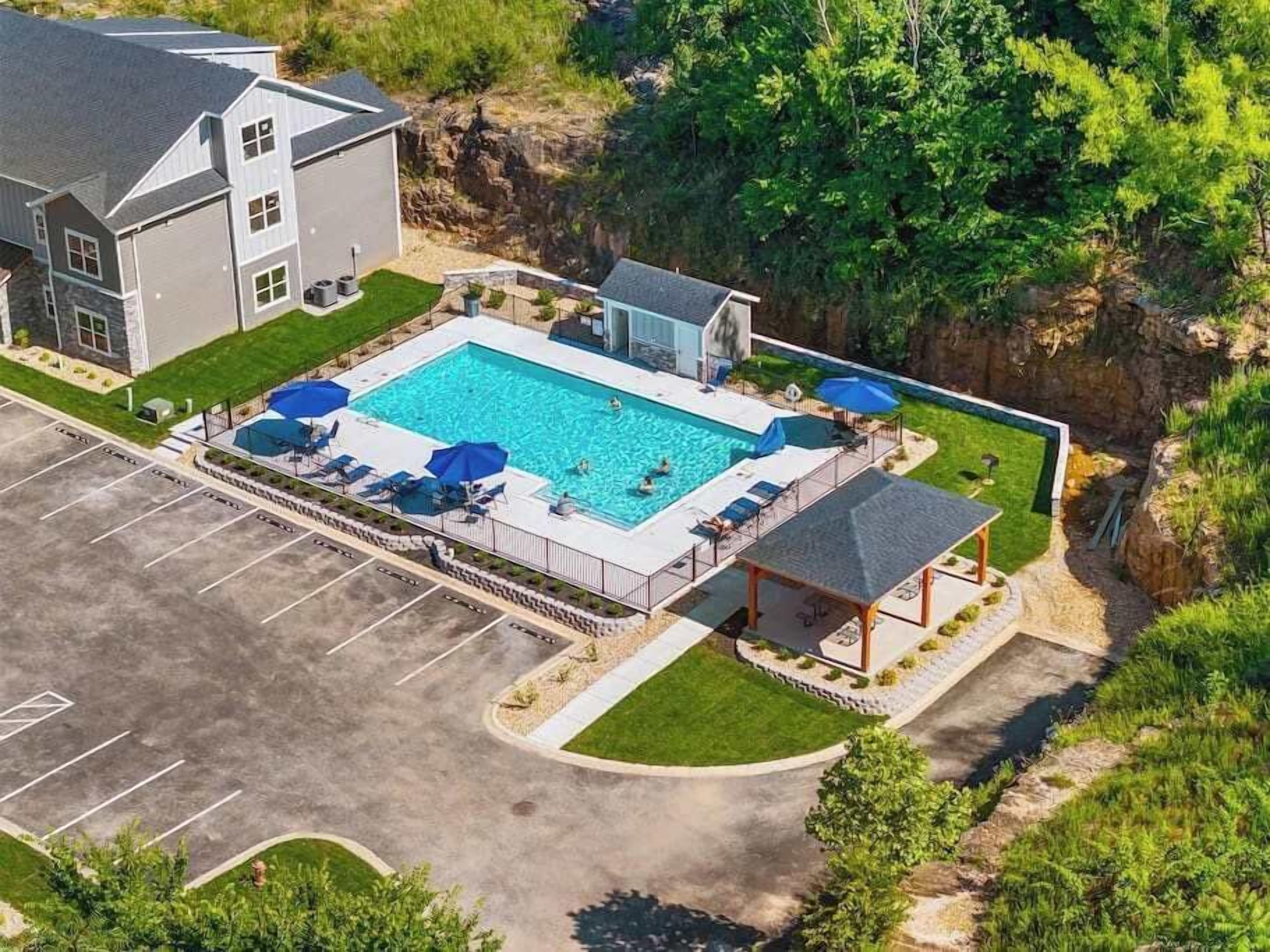 A swimming pool with lounge chairs and umbrellas, surrounded by a parking lot and a building.