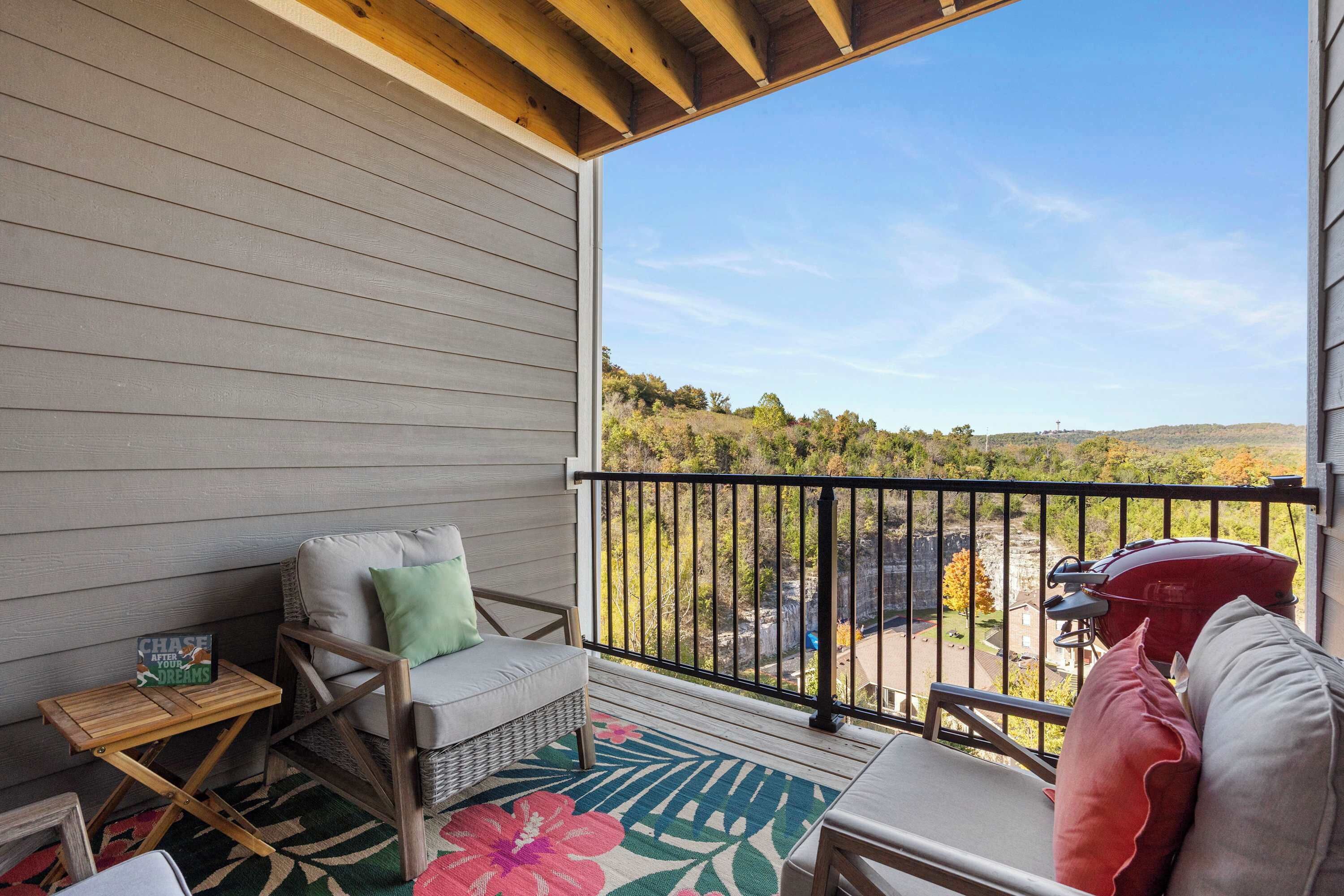 A covered balcony with a view of a hilly landscape, a wicker chair, a small table with a sign, and a barbecue grill.