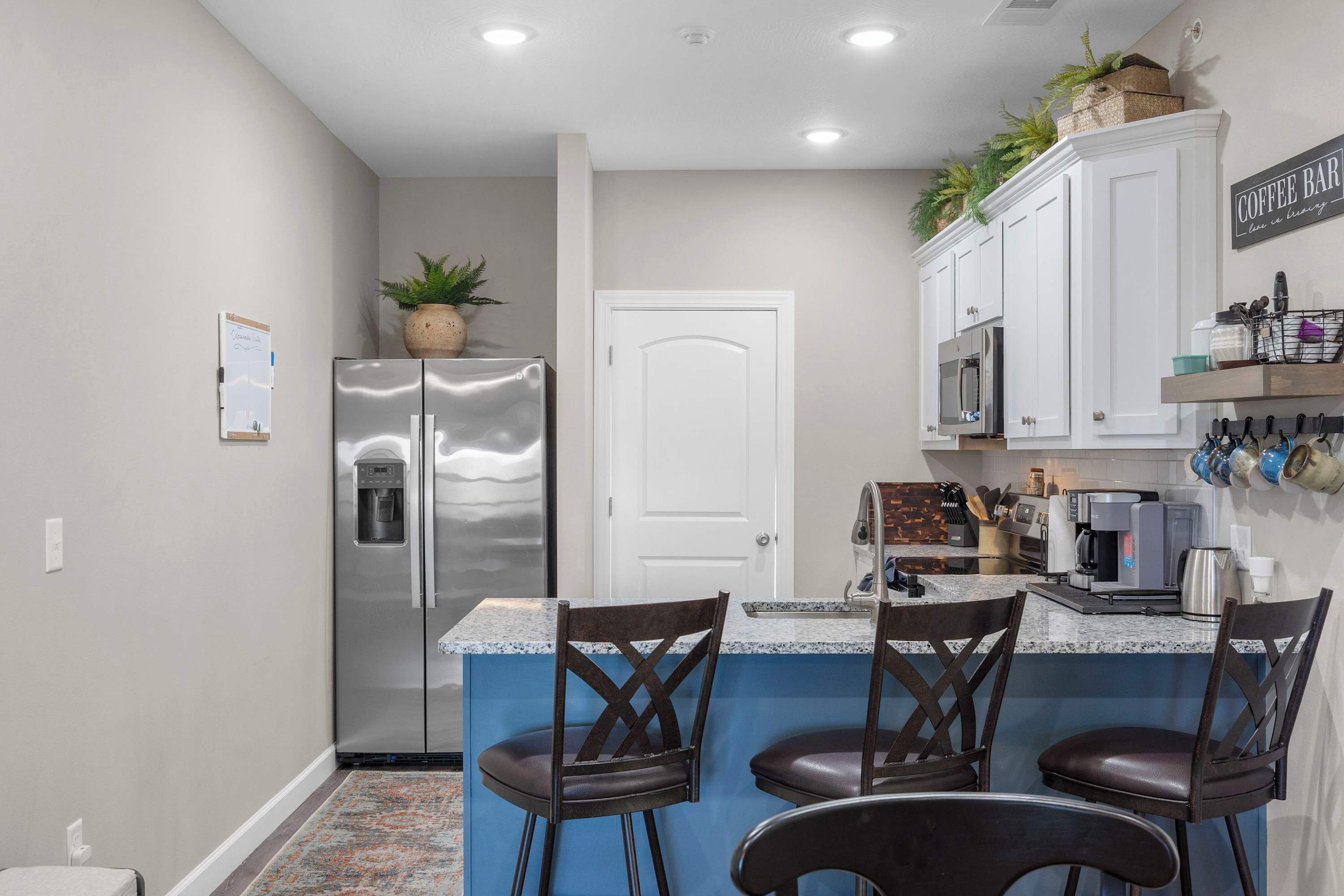 A modern kitchen with a stainless steel refrigerator, a built-in microwave, and a coffee bar. There are three dark brown chairs around a blue island counter.