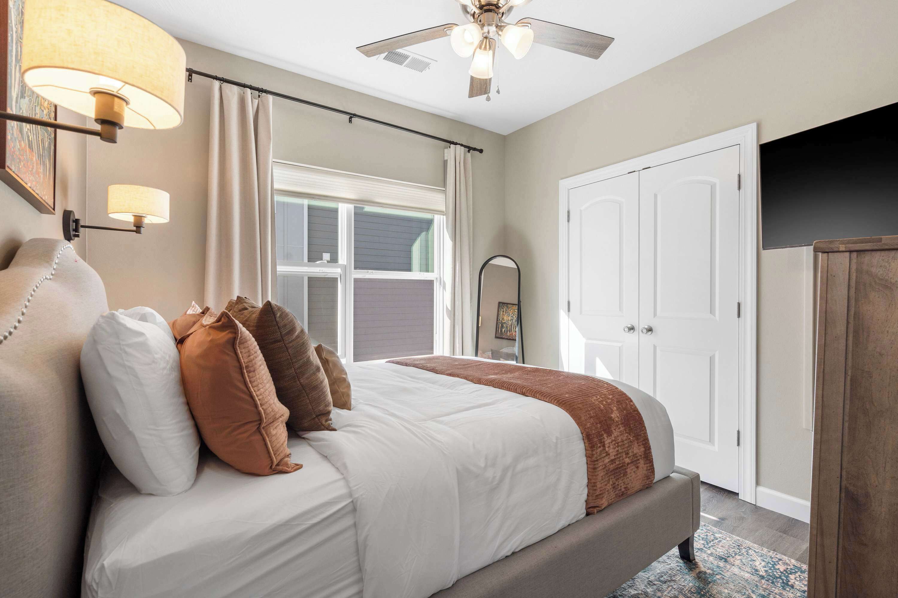 A bedroom with a bed, a ceiling fan, a window with curtains, a door, and a mirror.