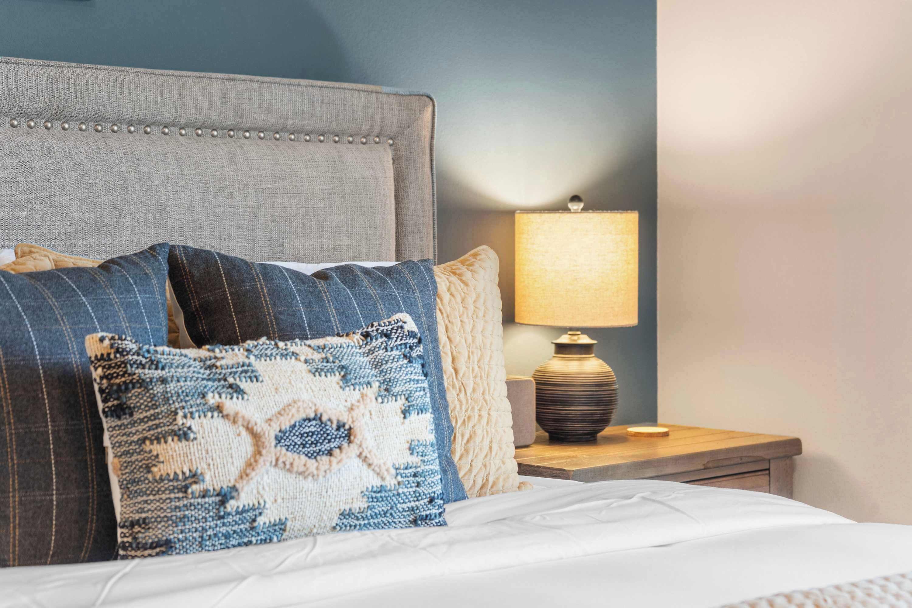 Een keurig opgemaakt bed met een grijs gestoffeerd hoofdbord, een bedlampje en een houten nachtkastje.