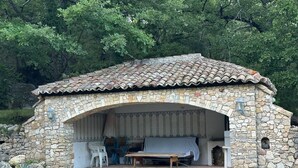 Un bâtiment en pierre avec un toit en tuiles, un espace de repos couvert avec une table et des chaises, et un mur de pierre qui entoure la structure.
