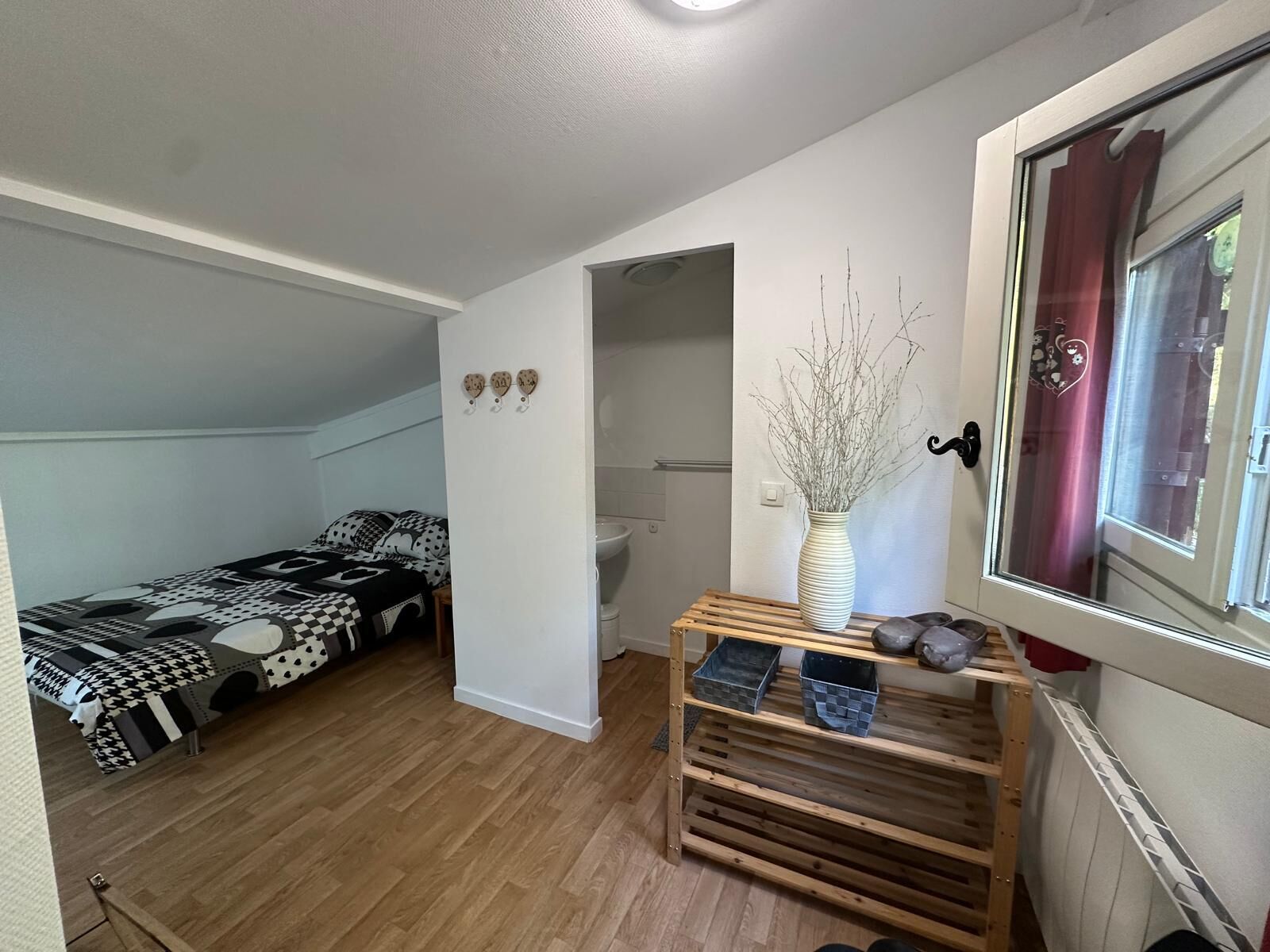 A small, well-lit room with a bed, a wooden shelf with storage boxes, and a window with curtains.