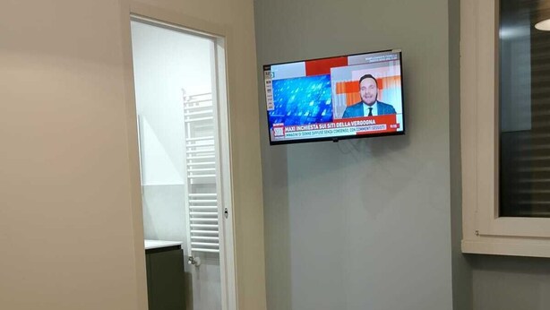 Hotellihuoneessa on seinälle asennettu televisio, jossa näkyy uutiskanava, sängyllä on rullalle taiteltuja pyyhkeitä ja ikkunassa on sälekaihtimet.