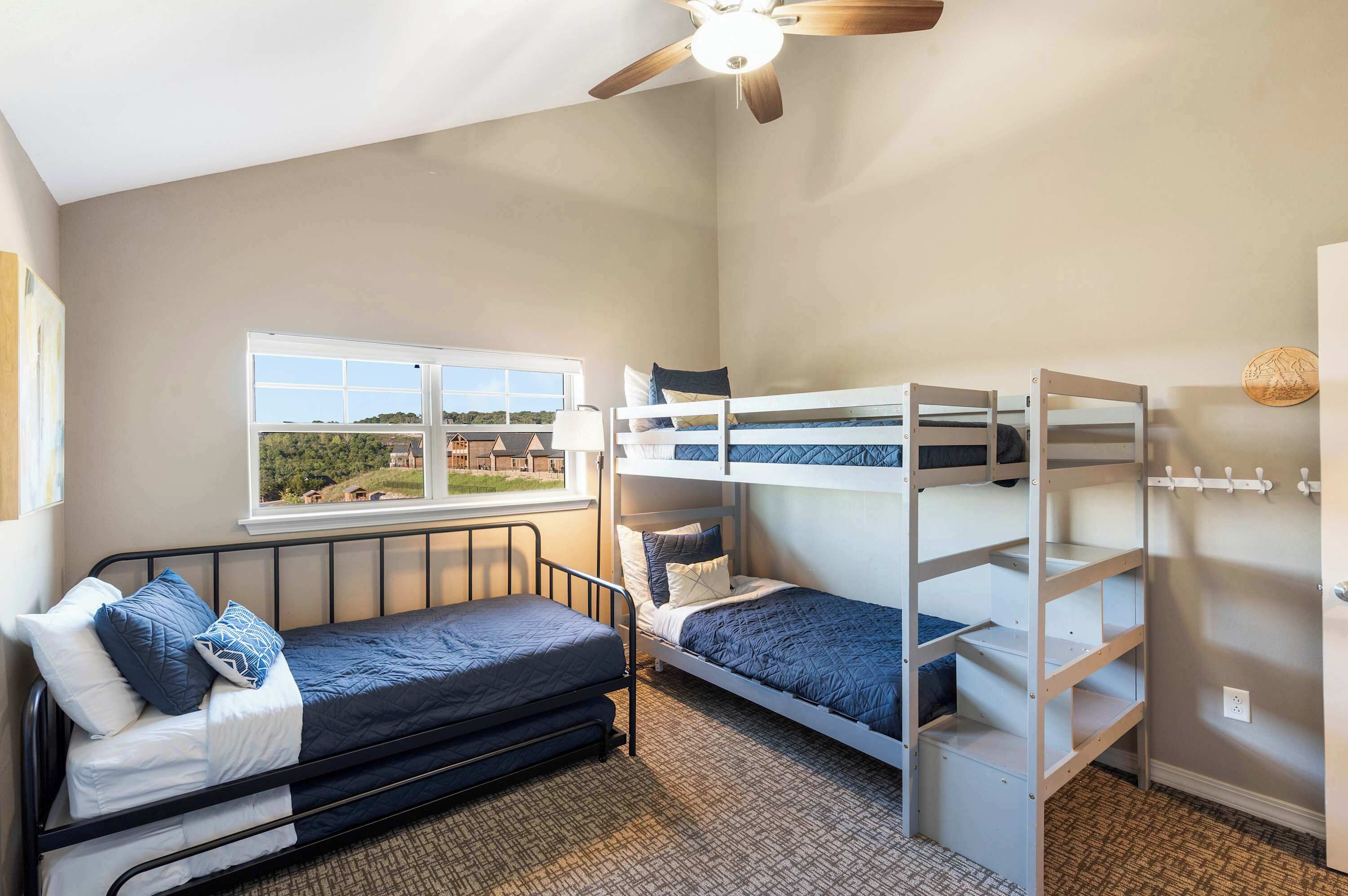A bedroom with a bunk bed, a single bed, a window with a view, a ceiling fan, and a wall-mounted coat rack.