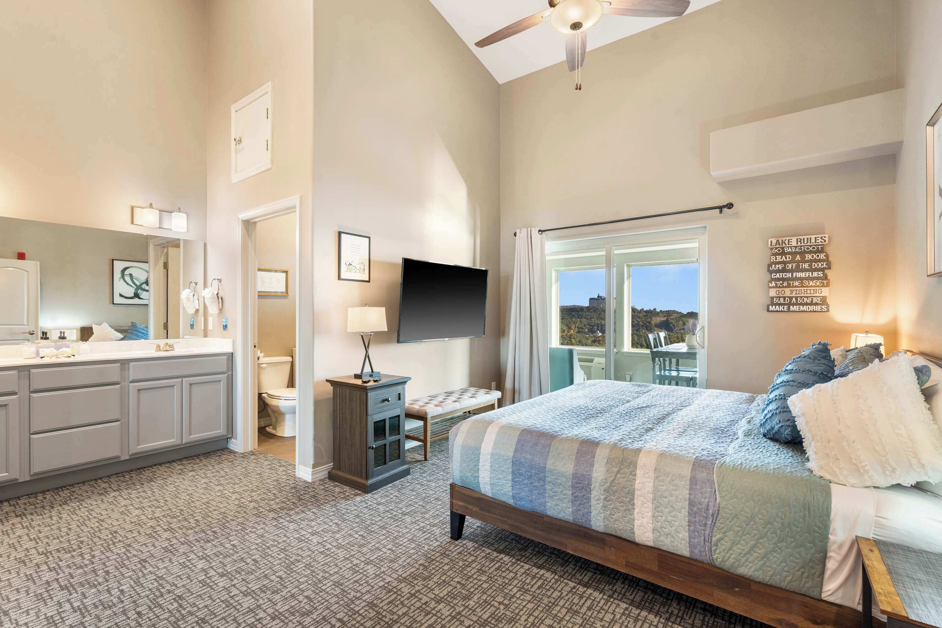 A bedroom with a large bed, a vanity with a mirror, a flat-screen TV, and a view of the outdoors.