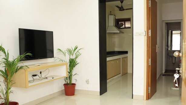 A spacious living area with a flat-screen TV on a wooden stand, two potted plants, a ceiling fan, and a kitchen visible through an open doorway. - Aaru Residency (Coimbatore)