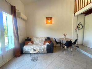 A modern living room with a sofa, a round coffee table, and a dining table with chairs. There is a wall-mounted light fixture above the sofa and a floor lamp near the dining area.