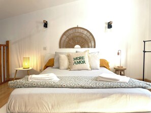A neatly made bed with a patterned bedspread, two pillows, and a decorative wall hanging. There are two bedside tables with lamps, a wooden chair, and a tall floor lamp.