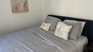 Una cama bien hecha con ropa de cama a rayas y varios almohadones, un cabecero oscuro y una pared minimalista con una sola obra de arte abstracta.