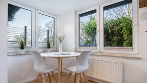 Ein modernes Zimmer mit einem runden Tisch, zwei weißen Stühlen, einem weißen Heizkörper und großen Fenstern mit Blick auf Bäume und ein Gebäude.